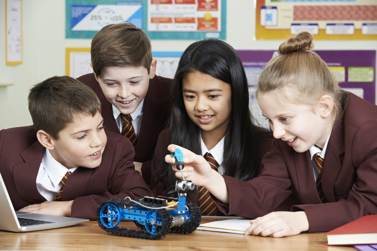 Four school students in school uniform sit around a desk as they experiment with a model robot and a laptop. 