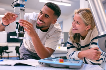 Two workers sit at a desk operating machinery. One is holding the apparatus while the other smiles.