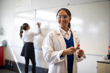 Two young people in a classroom. One is in the foreground holding a scientific model. The other writes on the whiteboard in the background.
