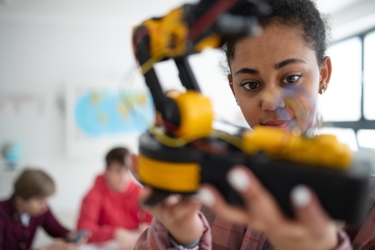 Young person holding a robot close to the camera, examining it. They are in a classroom with other students in the background.