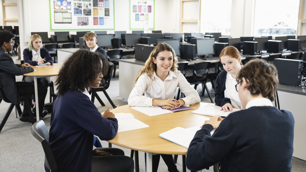 Two groups of secondary school students sitting and working around two tables in a classroom setting. They have work books on the desks and are talking.