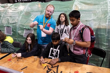 A volunteer at The Big Bang Fair demonstrates a task to four young people. Two of these people are sat at at table as they work on the task while the other two stand. All students are wearing lanyards.