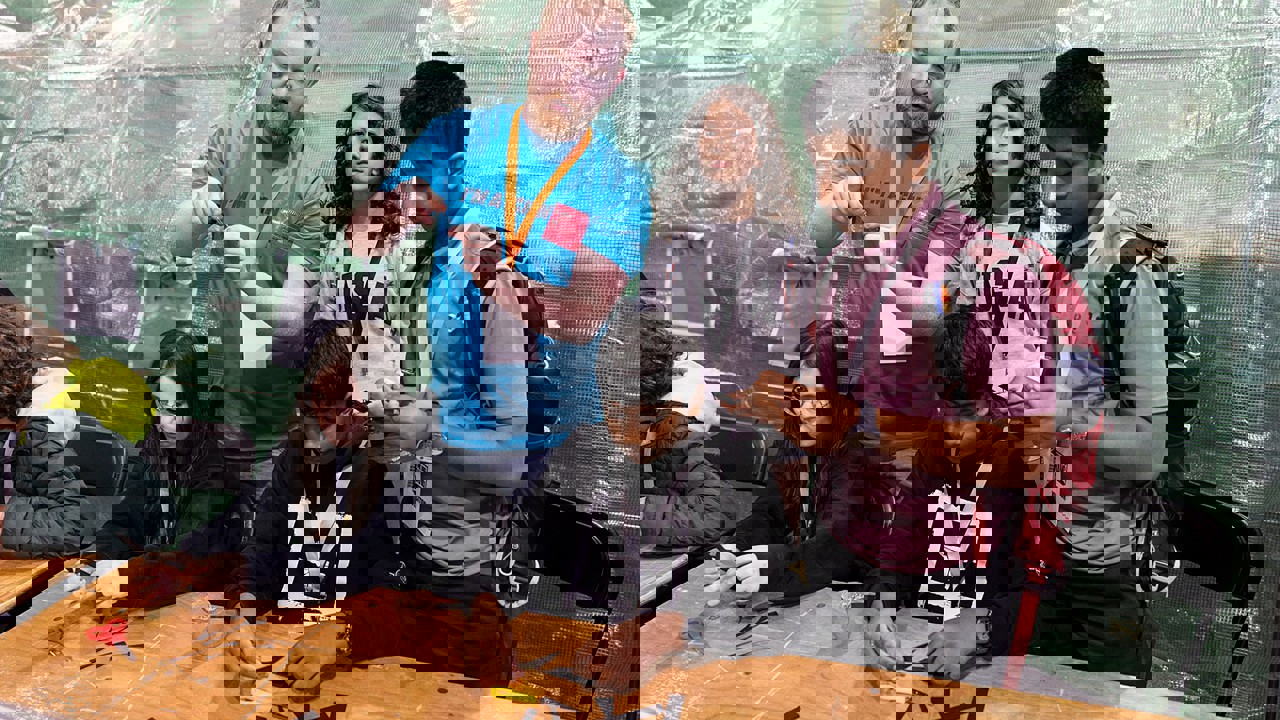 A volunteer at The Big Bang Fair demonstrates a task to four young people. Two of these people are sat at at table as they work on the task while the other two stand. All students are wearing lanyards.