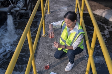 An engineer crouches on a footbridge over a body of water. They have beakers with chemicals. They wear high visibility outerwear. 