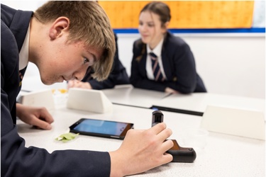 A school classroom. The focus is on one young person who is looking intently at a microscope as other members of the class appear in the background. 