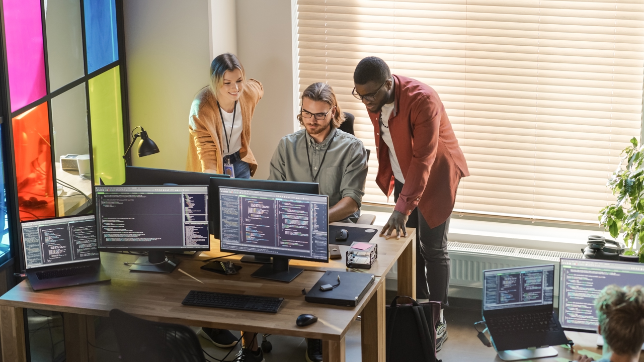 Three people in a colourful office environment. One sits at their desk while the others stand either side. They all look at the screen. 