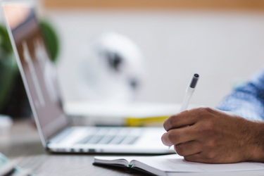 A close-up of a laptop and notepad. A hand makes notes in the notepad with a pen