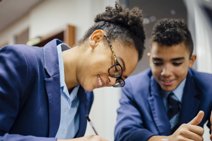 Secondary school students in a classroom at a desk working. One writes as the others look on.