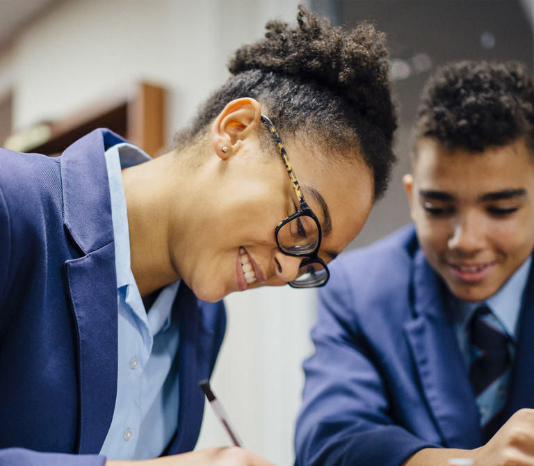 Secondary school students in a classroom at a desk working. One writes as the others look on.