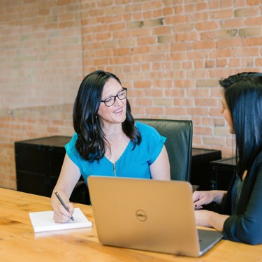 Two people sit at a large desk at work. They face each other. One has a pen and notepad, one has a laptop.