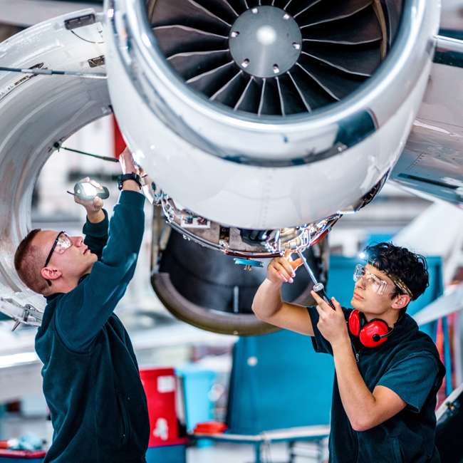 Two engineers working on an aircraft. They have protective wear and tools and are underneath a propeller. 