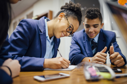 Secondary school students in a classroom at a desk working. One writes as the others look on.