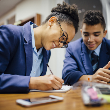 Secondary school students in a classroom at a desk working. One writes as the others look on.