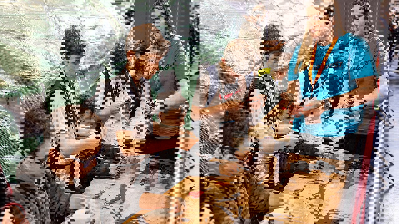 A selection of young people are working to complete a task with materials such as scissors, card, recyclable pots and paper clips. Some are sitting, some are standing. A volunteer watches on to support.