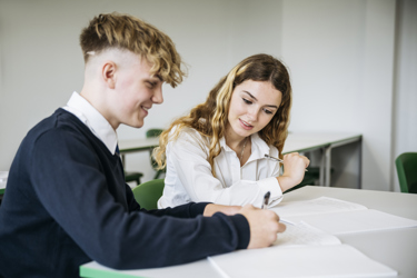 Two secondary school pupils sit at a desk in a classroom working together. One is writing in their text book. 