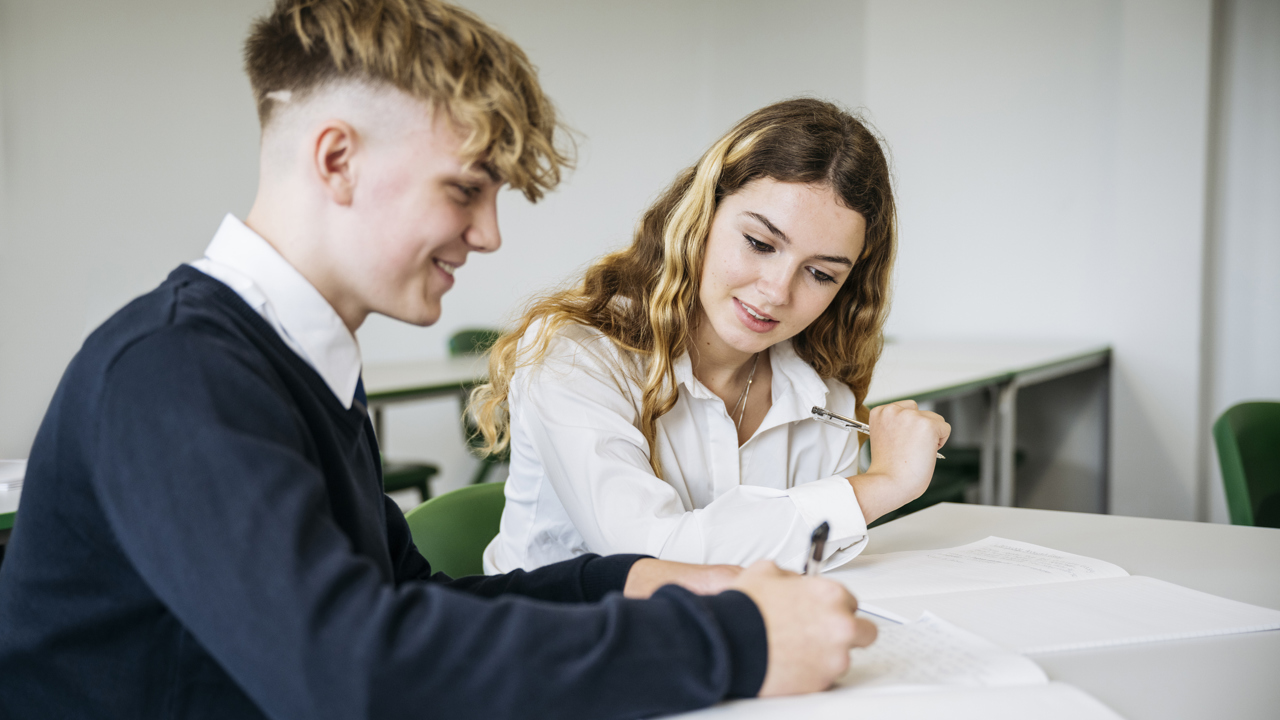 Two secondary school pupils sit at a desk in a classroom working together. One is writing in their text book. 