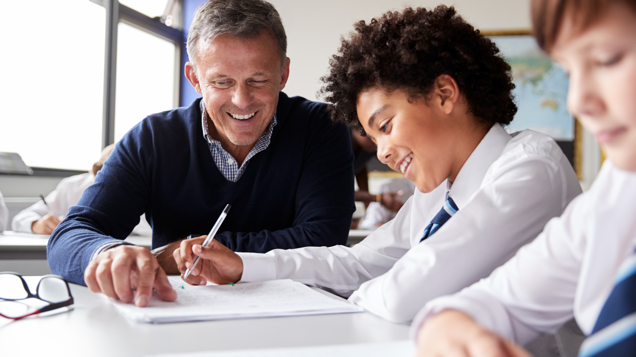 In a classroom, a teacher sits with two pupils as they work at their desks