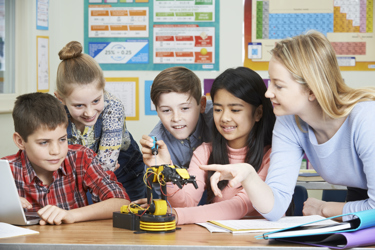 Four primary school pupils and one teacher sit at a desk watching one of the pupils as they conduct an experiment with a model robot and laptop.