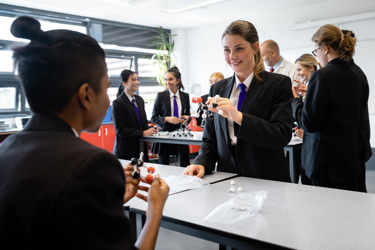 Several secondary school pupils and a teacher in a classroom partaking in a science lesson. Some students are doing experiments while others watch.