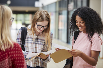 Three secondary school students in a school corridor smile as they each look at the contents of a letter.