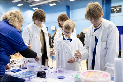 A school science fair setting. Four young people visit a stand and are conducting an experiment supported by a grown up. They all wear protective clothing and goggles. They work with electronic pipettes and liquids in beakers and flasks.