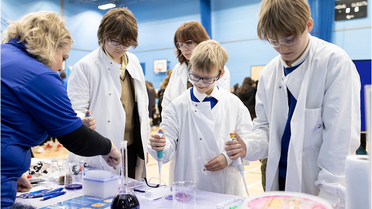 A school science fair setting. Four young people visit a stand and are conducting an experiment supported by a grown up. They all wear protective clothing and goggles. They work with electronic pipettes and liquids in beakers and flasks.