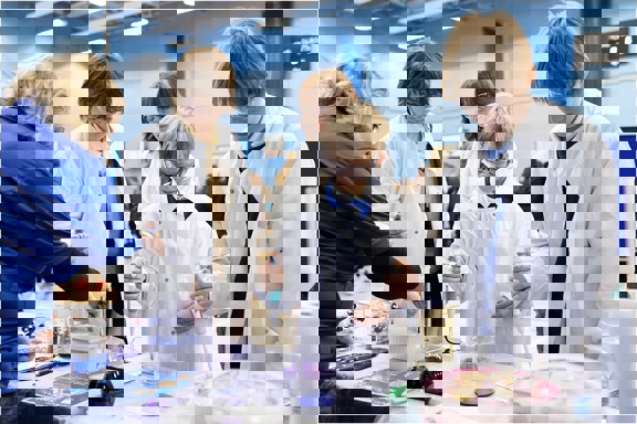 A school science fair setting. Four young people visit a stand and are conducting an experiment supported by a grown up. They all wear protective clothing and goggles. They work with electronic pipettes and liquids in beakers and flasks.