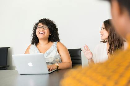 Three people sit around a table in an office meeting room setting. One has their back to the camera. One is at their laptop. One has their hand raised.