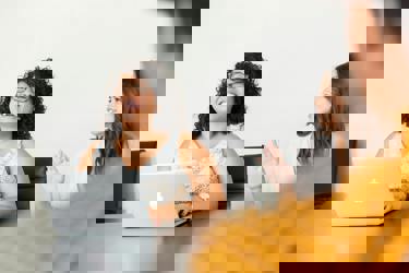 Three people sit around a table in an office meeting room setting. One has their back to the camera. One is at their laptop. One has their hand raised.