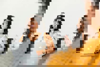 Three people sit around a table in an office meeting room setting. One has their back to the camera. One is at their laptop. One has their hand raised.