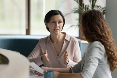 Two adults sit at a table at work. They face each other in discussion. One is holding a piece of paper as they look at their colleague. 