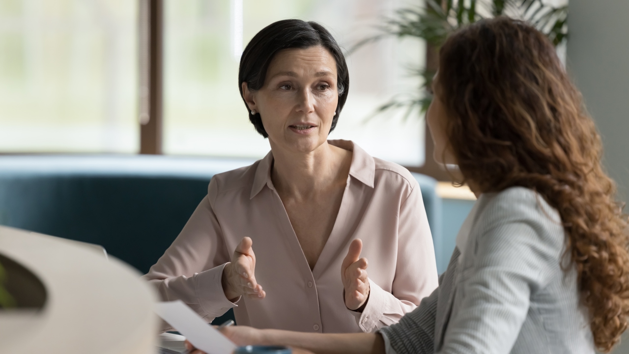 Two adults sit at a table at work. They face each other in discussion. One is holding a piece of paper as they look at their colleague. 