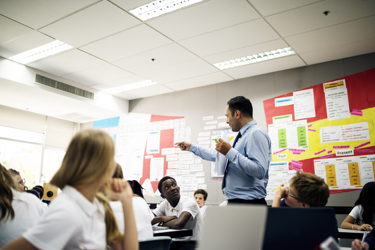 A busy classroom with a teacher standing talking and pointing while students sit and listen. There is a colourful noticeboard in the room.