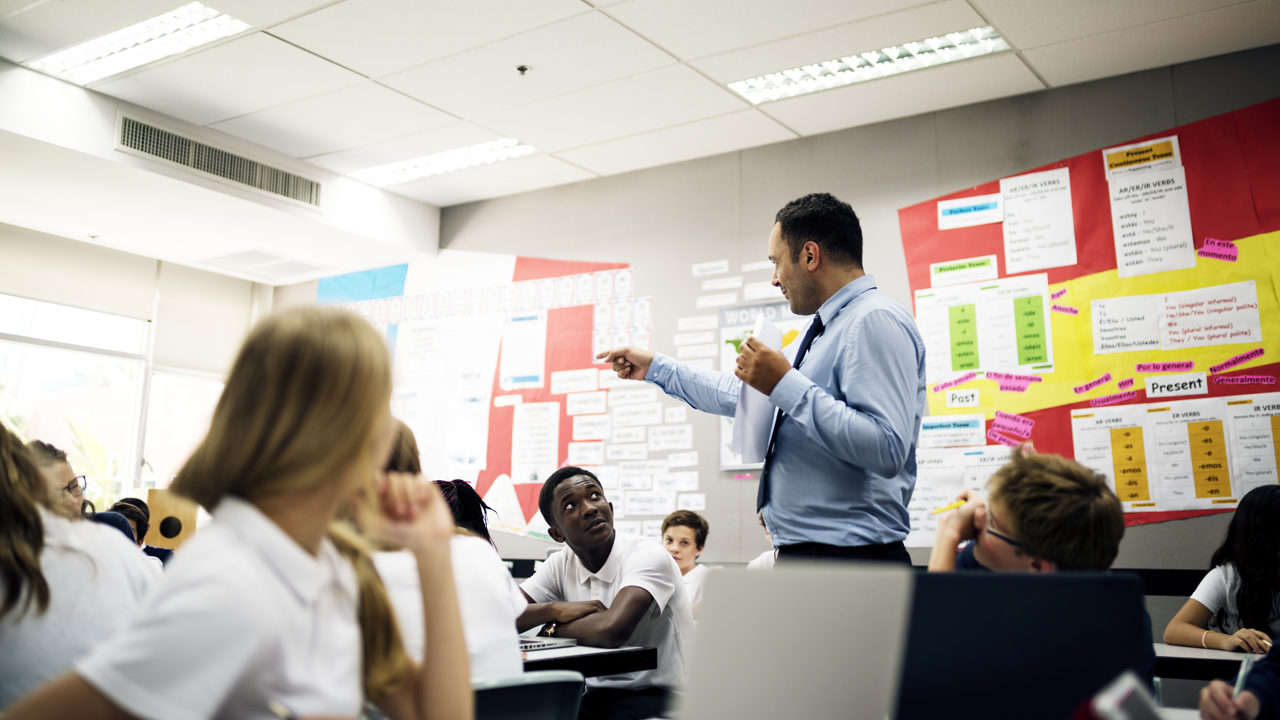 A busy classroom with a teacher standing talking and pointing while students sit and listen. There is a colourful noticeboard in the room.