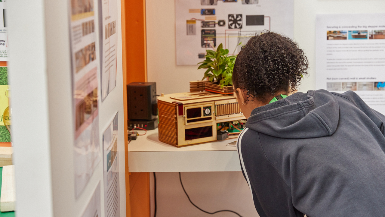 A booth featuring display boards to explain how a scientific competition entry evolved. A young person with their back to the camera bends down and peers at the scale model on display.