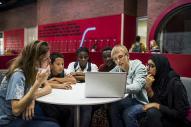 Six young people sit at a round table watching a laptop screen intently. 