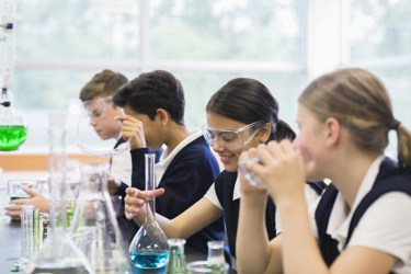 Four secondary school pupils working in a science lesson. They are all wearing protective goggles as they work with scientific equipment such as beakers, flasks and coloured liquids. 