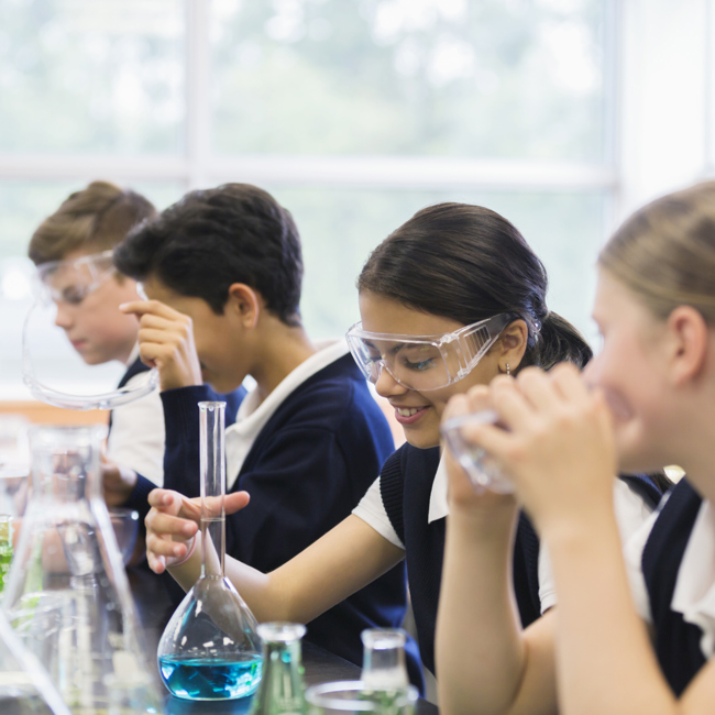 Four secondary school pupils working in a science lesson. They are all wearing protective goggles as they work with scientific equipment such as beakers, flasks and coloured liquids. 