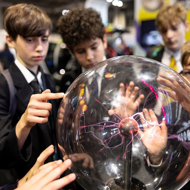 Six hands touch a plasma ball on display at The Big Bang Fair. Four young people are watching the effects.