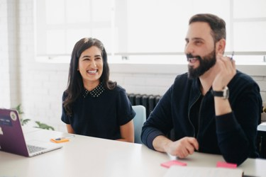 Two adults in an office meeting room setting. One has a laptop the other is talking. There are post-it notes on the desk alongside a laptop. 