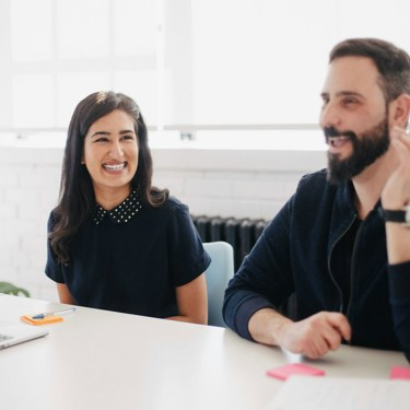 Two adults in an office meeting room setting. One has a laptop the other is talking. There are post-it notes on the desk alongside a laptop. 