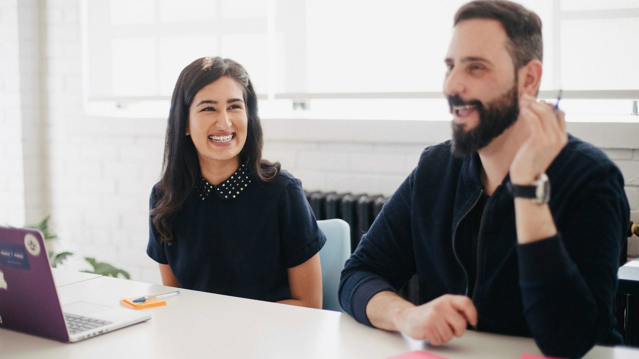 Two adults in an office meeting room setting. One has a laptop the other is talking. There are post-it notes on the desk alongside a laptop. 