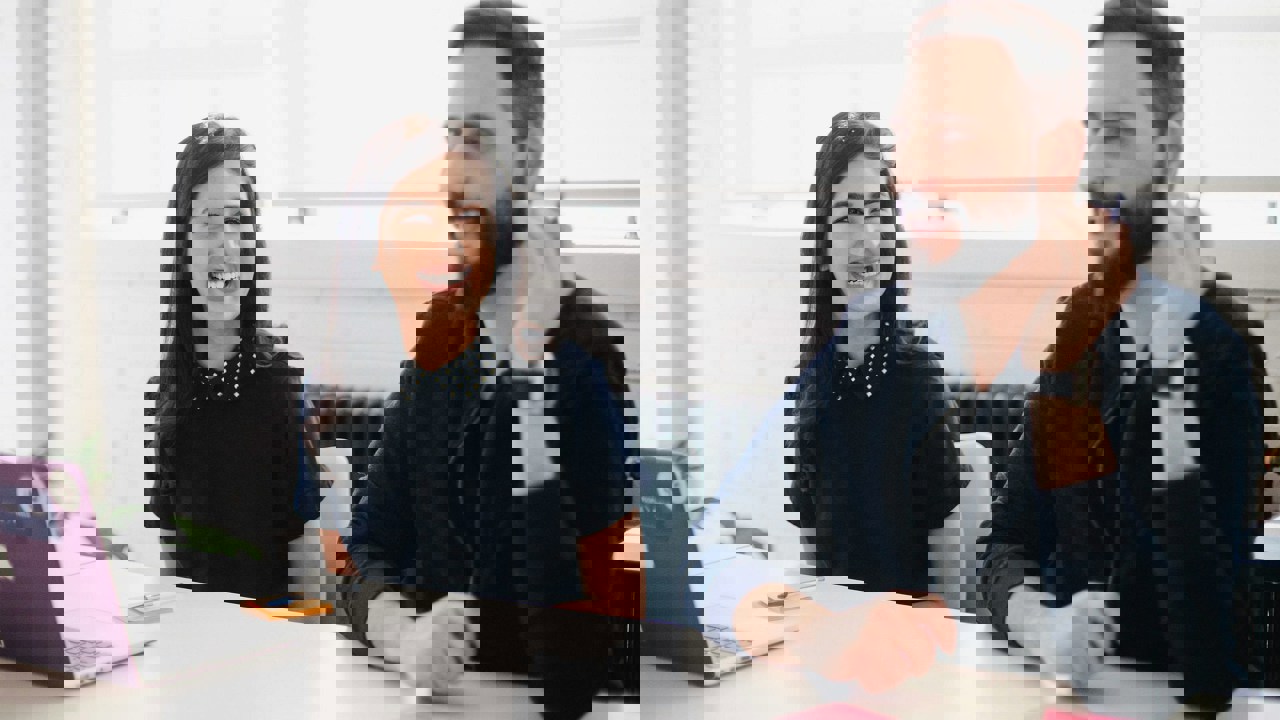 Two adults in an office meeting room setting. One has a laptop the other is talking. There are post-it notes on the desk alongside a laptop.