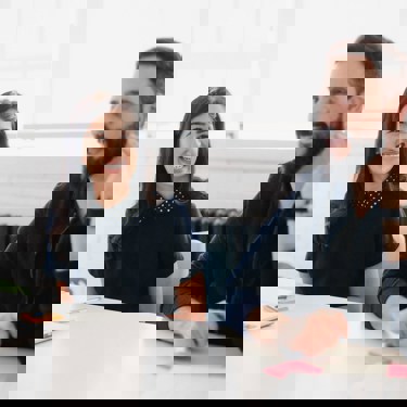 Two adults in an office meeting room setting. One has a laptop the other is talking. There are post-it notes on the desk alongside a laptop.