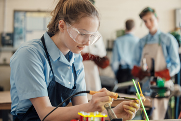 A secondary school student is in a classroom in a practical lesson. They are wearing protective goggles and apron as they work with electronic equipment, focusing on the materials on their workbench.