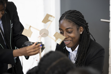 Close up shot of a group of young people building a model of a wind turbine in their science lesson. One young person is holding the model as the camera focuses on another smiling