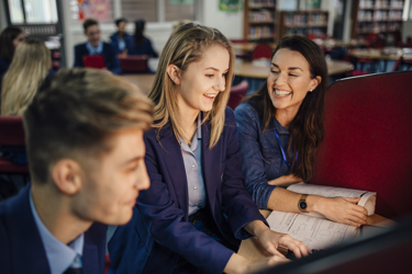 Secondary school students in a school library setting are in front of a screen being supported by their teacher. They have workbooks on the desk. 