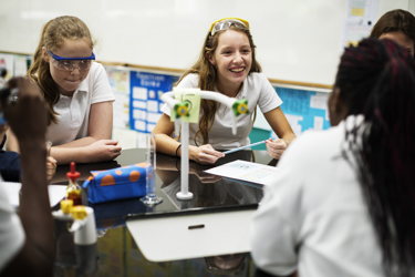 A group of secondary school students in a school science lab sit around a work bench with their pens and work book. There is scientific equipment on the surface too. The students are wearing protective goggles.