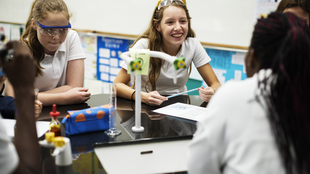 A group of secondary school students in a school science lab sit around a work bench with their pens and work book. There is scientific equipment on the surface too. The students are wearing protective goggles.