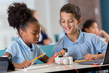 Primary school pupils in a classroom working with a laptop and a working model. One holds a pencil and presses a button on the model while the other watches and smiles.